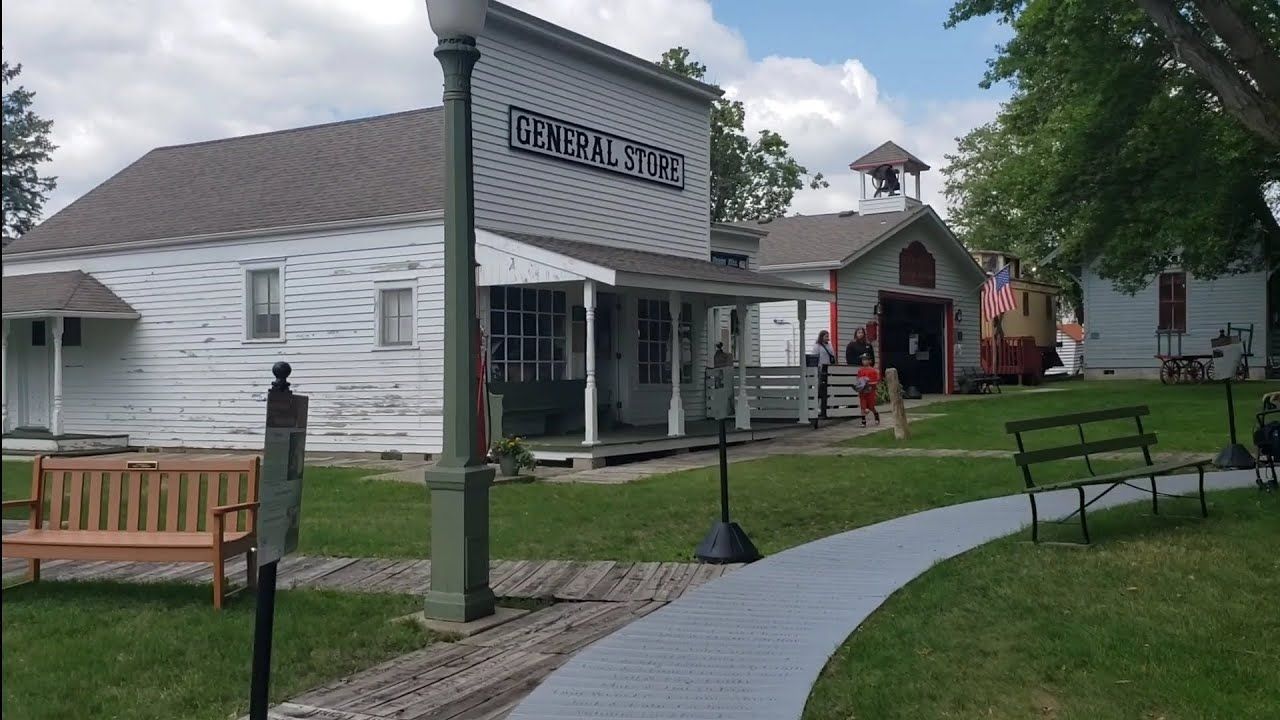 Old white buildings with a sign, a path, a lamp post, and a bench on grass.