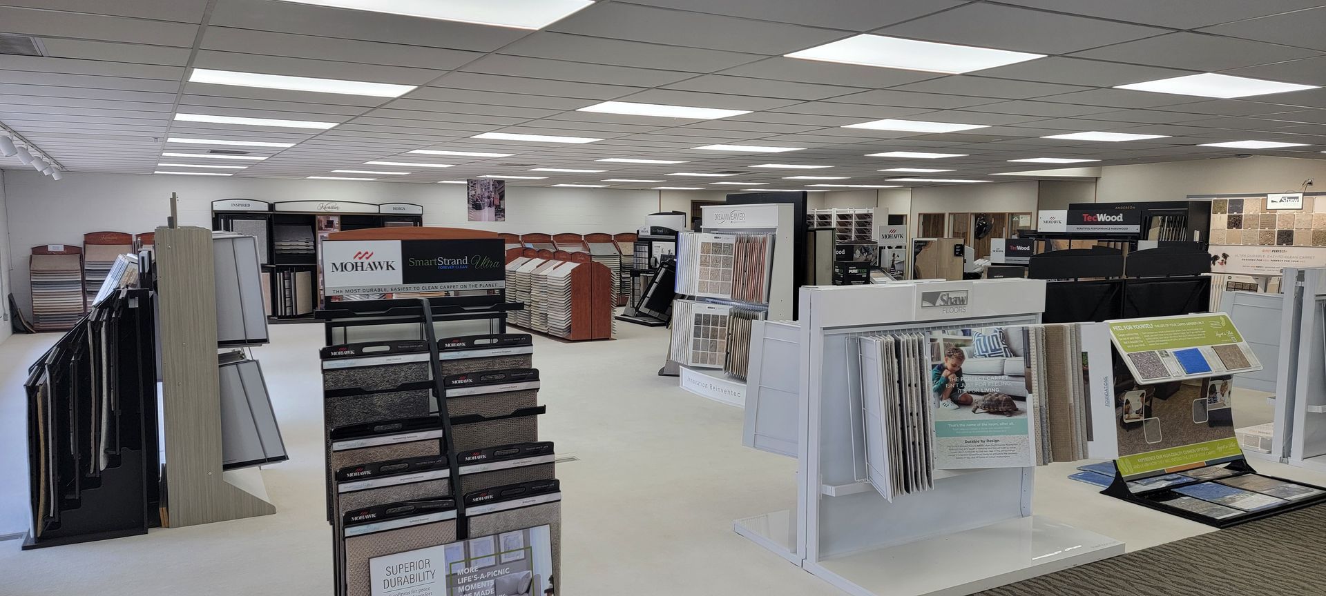 A retail showroom with flooring and display materials on tables and racks under bright overhead lights. A retail showroom with flooring and display materials on tables and racks under bright overhead lights.