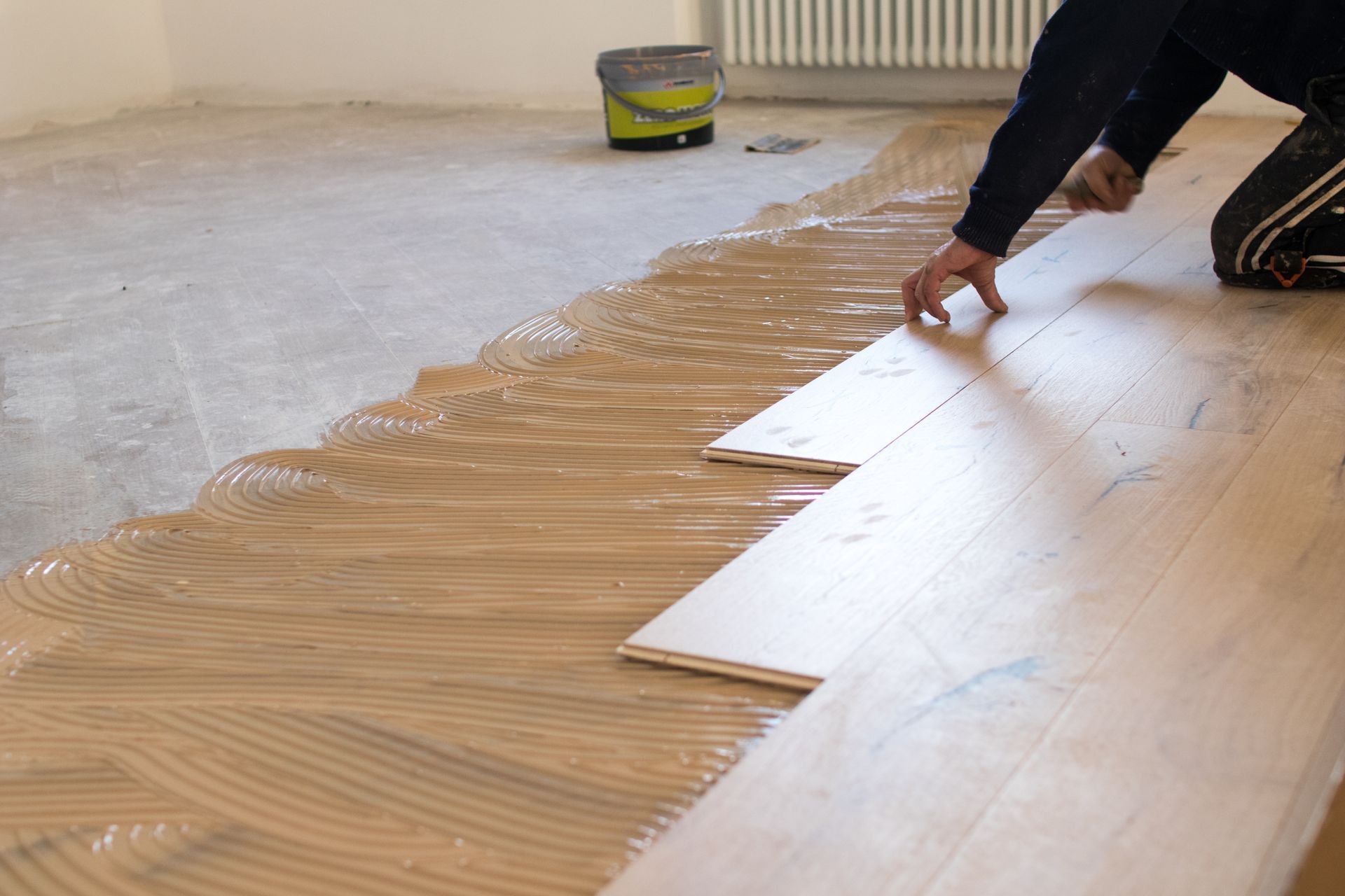 Person installing wood flooring, applying boards to a layer of glue on concrete floor in an interior setting. Person installing wood flooring, applying boards to a layer of glue on concrete floor in an interior setting.