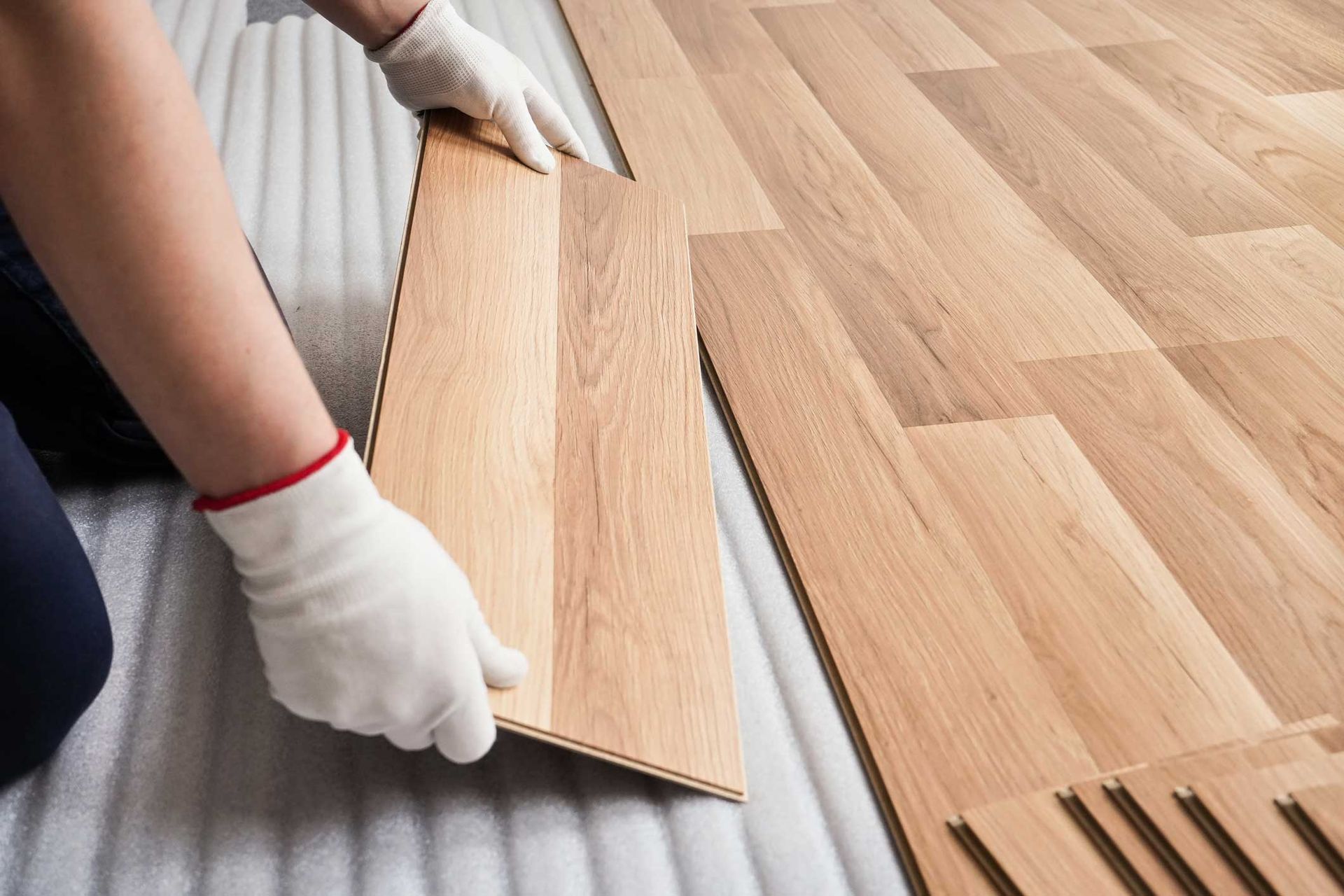 Hands wearing white gloves installing light wood flooring over underlayment. Hands wearing white gloves installing light wood flooring over underlayment.