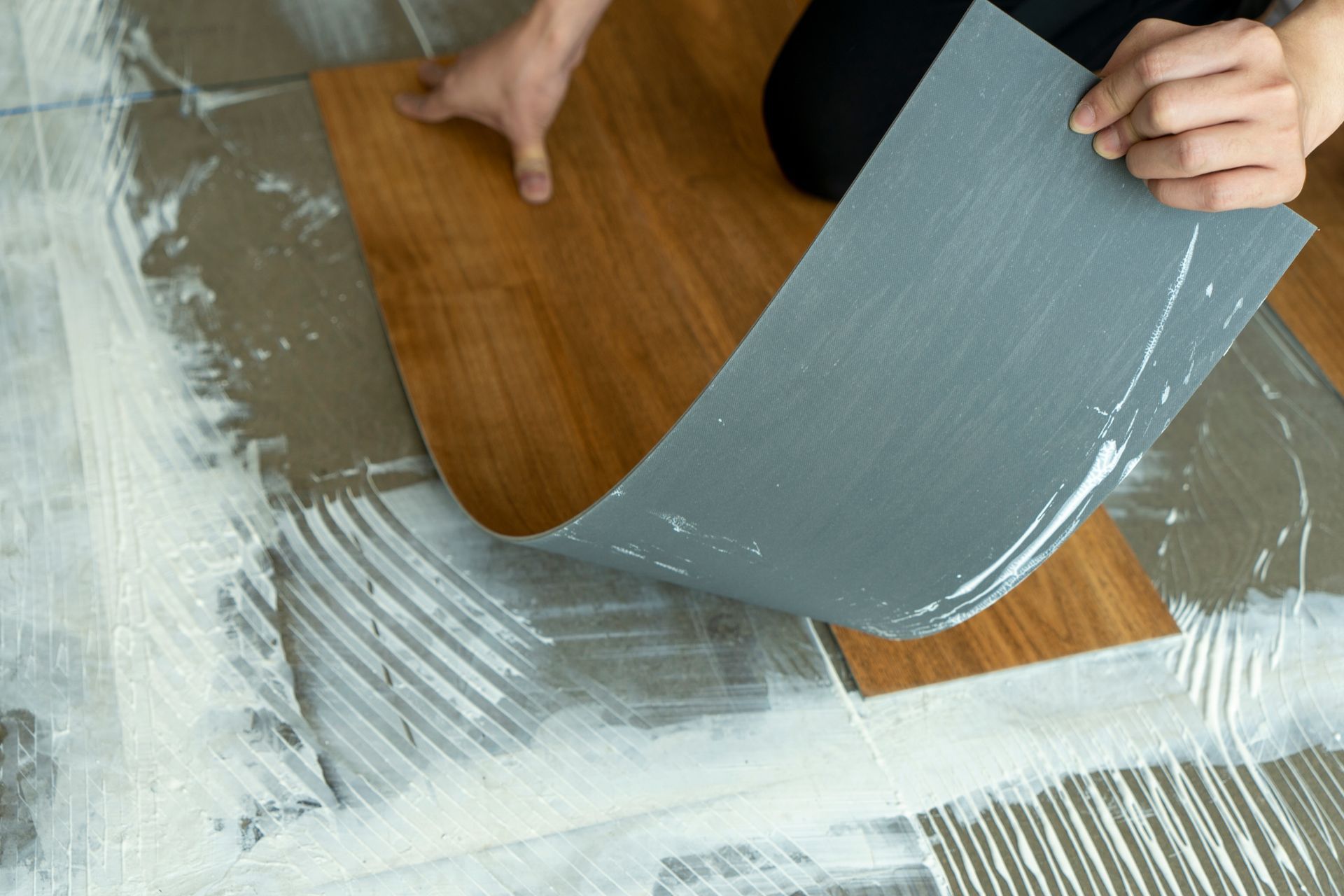 Person installing wood-look vinyl flooring, lifting a sheet of gray backing to apply. Person installing wood-look vinyl flooring, lifting a sheet of gray backing to apply.