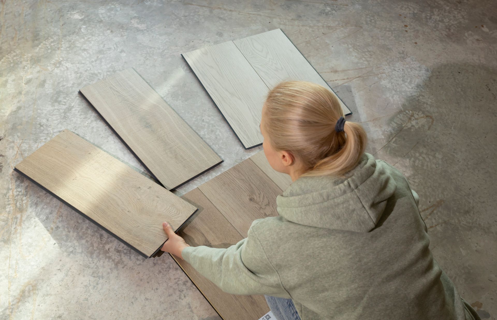 Person installing wood flooring with tools: hammer, level, tape measure. Woman on floor examining wood flooring samples.