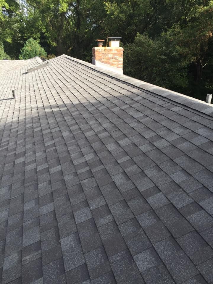 Gray shingle roof on a house with a brick chimney, surrounded by green trees.