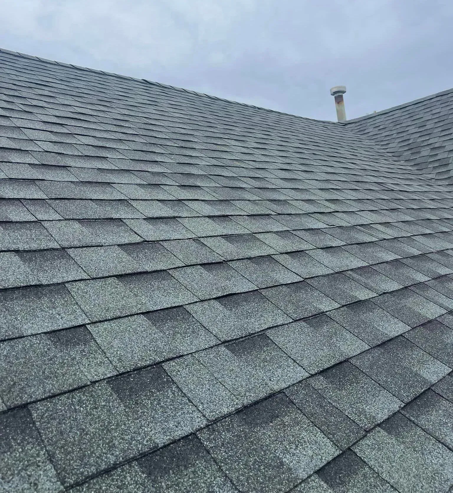 Gray asphalt shingle roof against a cloudy sky with a vent pipe visible.