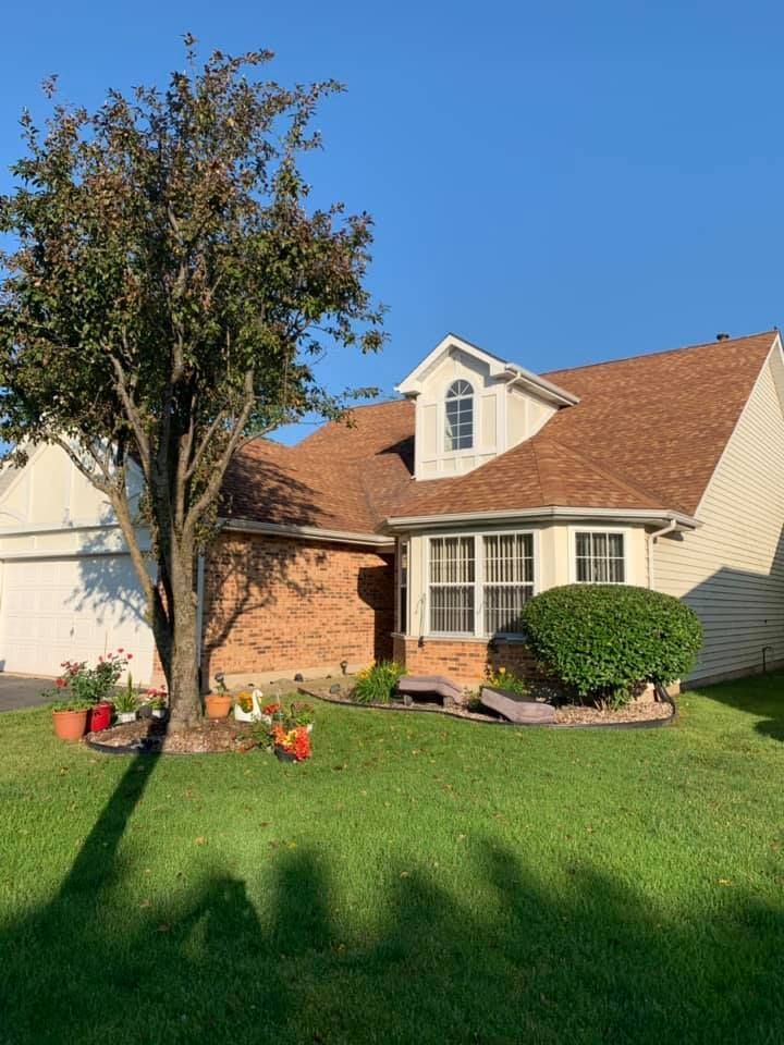 House with brown roof, brick and siding walls, green lawn, and a tree casting a shadow on the grass.