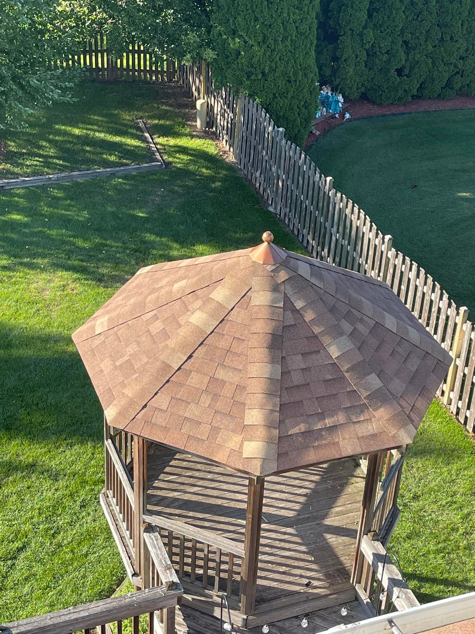 Gazebo with brown roof, surrounded by green grass, wooden fence, and trees.