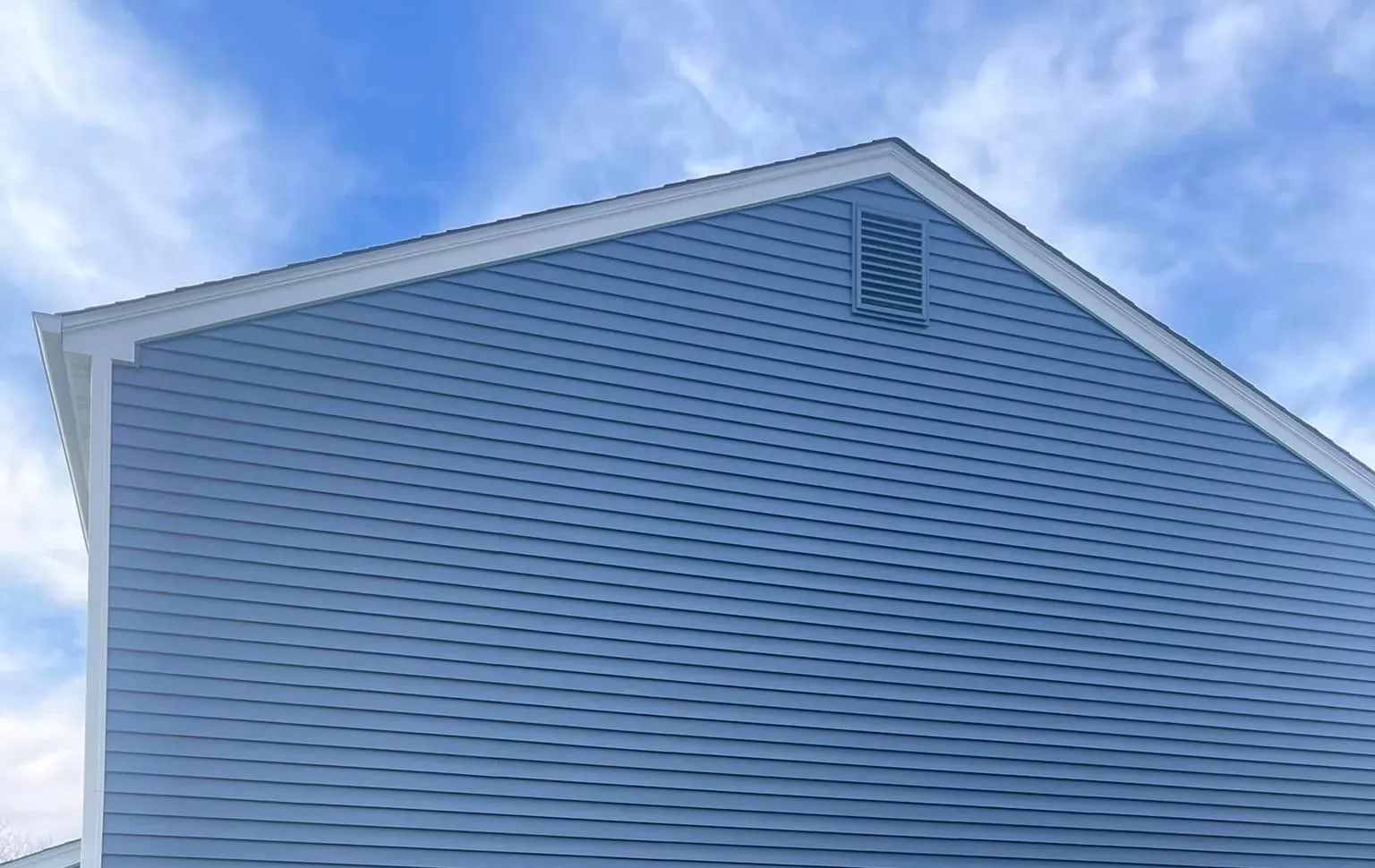 Blue siding on a house with a white trim and a vent in the blue sky background.