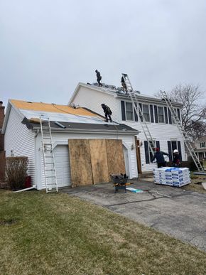 Roofers replacing shingles on a two-story house with ladders, plywood, and roofing supplies.