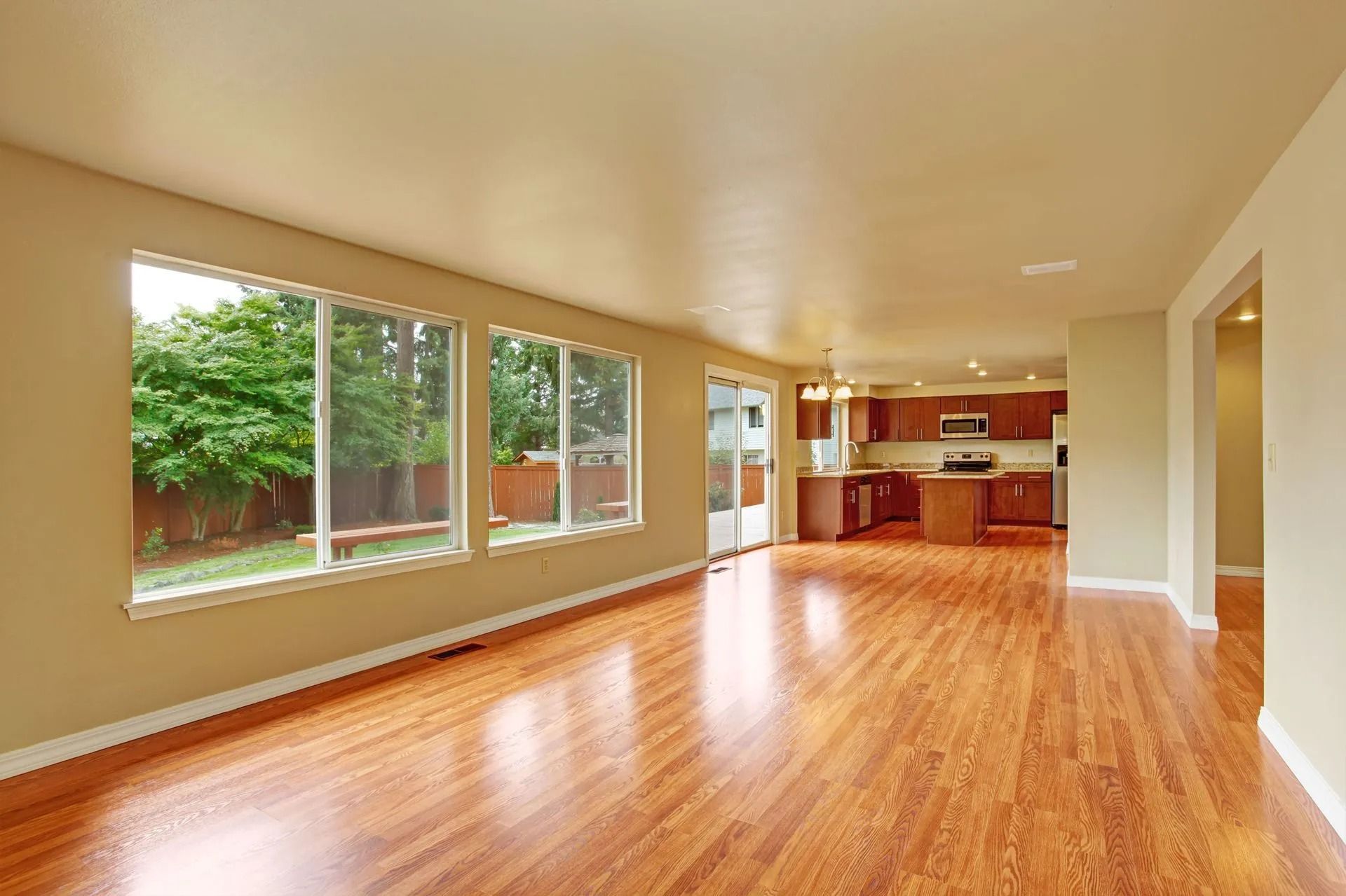 Empty living room with hardwood floor, large windows, and kitchen visible in the background.