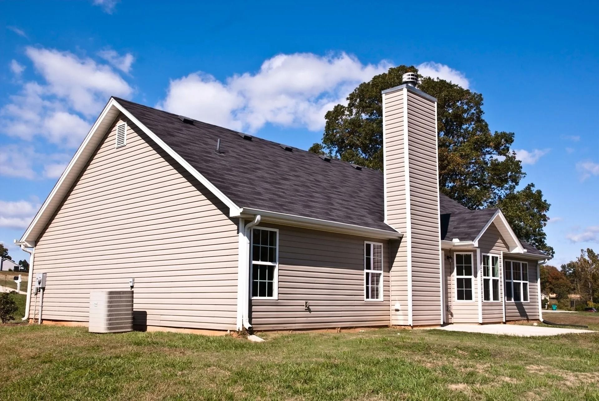 Beige house with dark roof, white trim, chimney, and HVAC unit on grassy lot under blue sky.