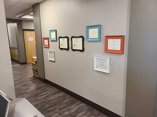 Hallway with framed certificates on the wall; beige walls, patterned carpet, and open doorways.