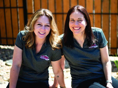 Two women smiling outdoors, wearing grey shirts with logo, wooden fence backdrop.
