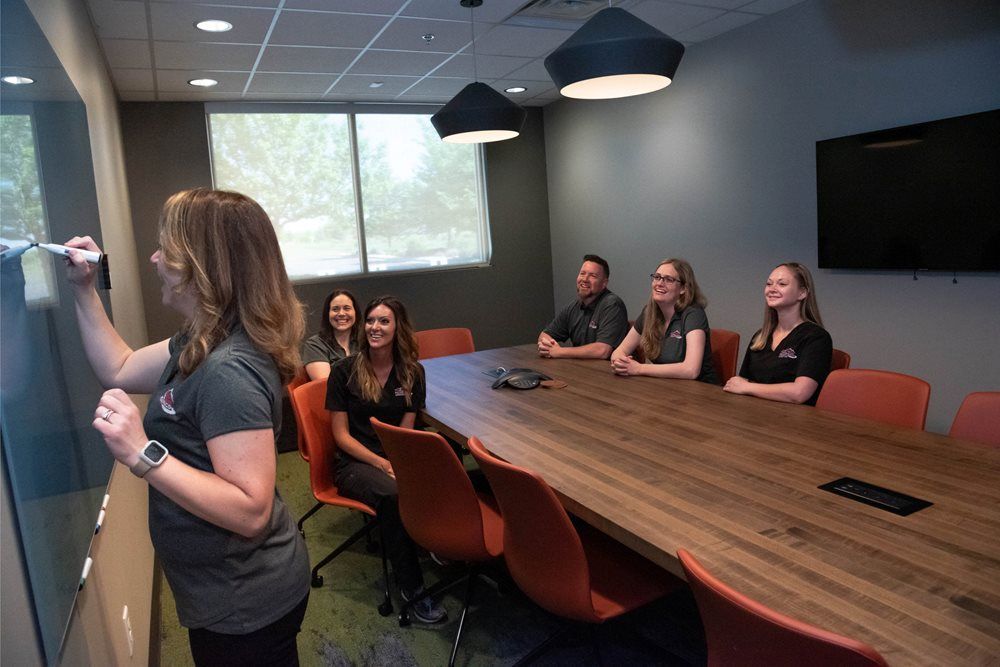 Woman writing on a whiteboard in a conference room with five people seated at a long table.