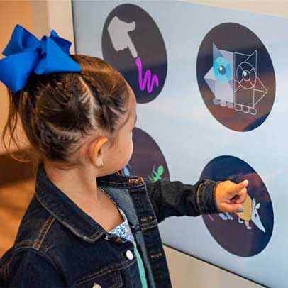 Girl pointing at a touch screen with animal icons at a museum. She wears a denim jacket and a blue bow.