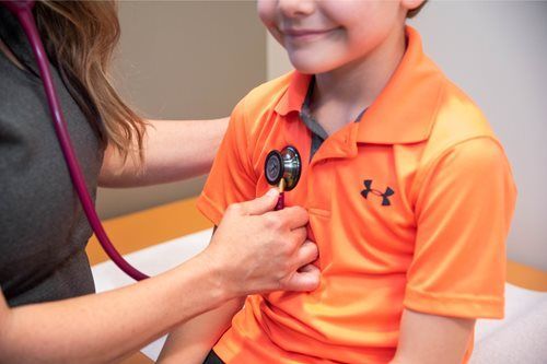Doctor listening to a child's chest with a stethoscope in a medical office setting. The child wears an orange shirt.