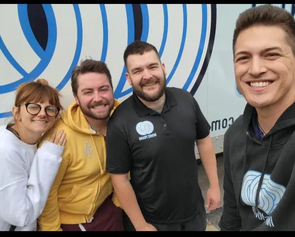 Four smiling people pose for a photo. They stand near a white and blue vehicle.