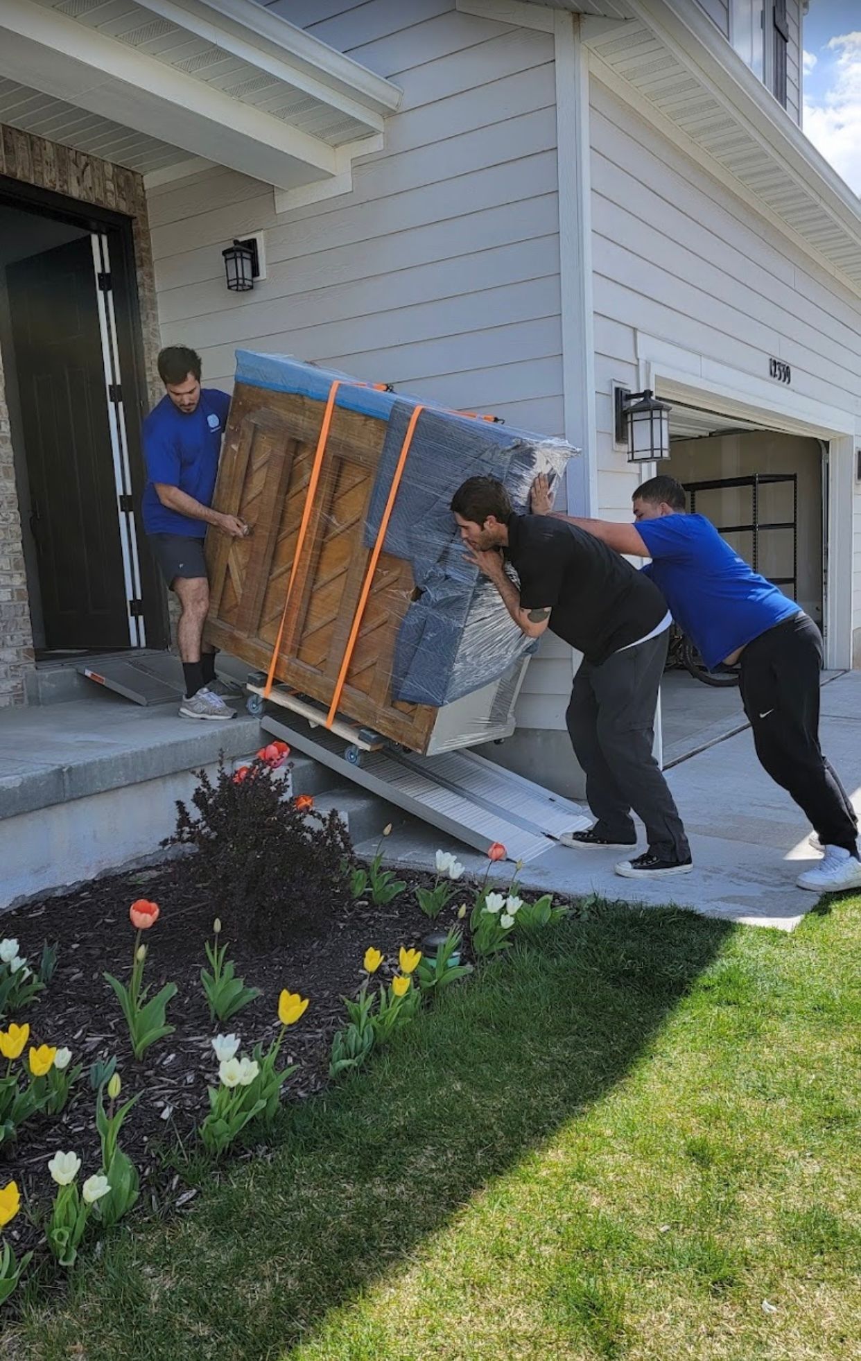 Three men moving a wrapped wooden cabinet on a ramp, near a house entrance.