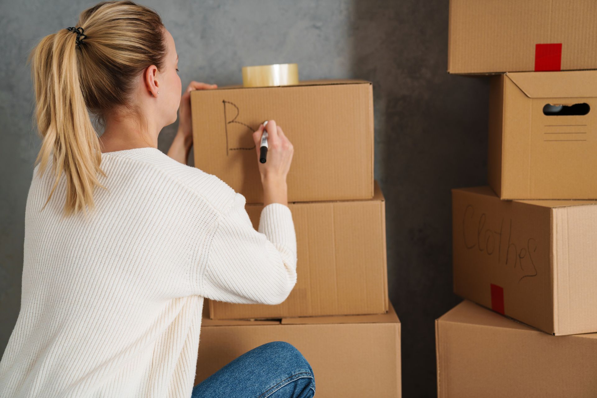 Woman writing on a cardboard box, taping it, stacks of boxes in a room.