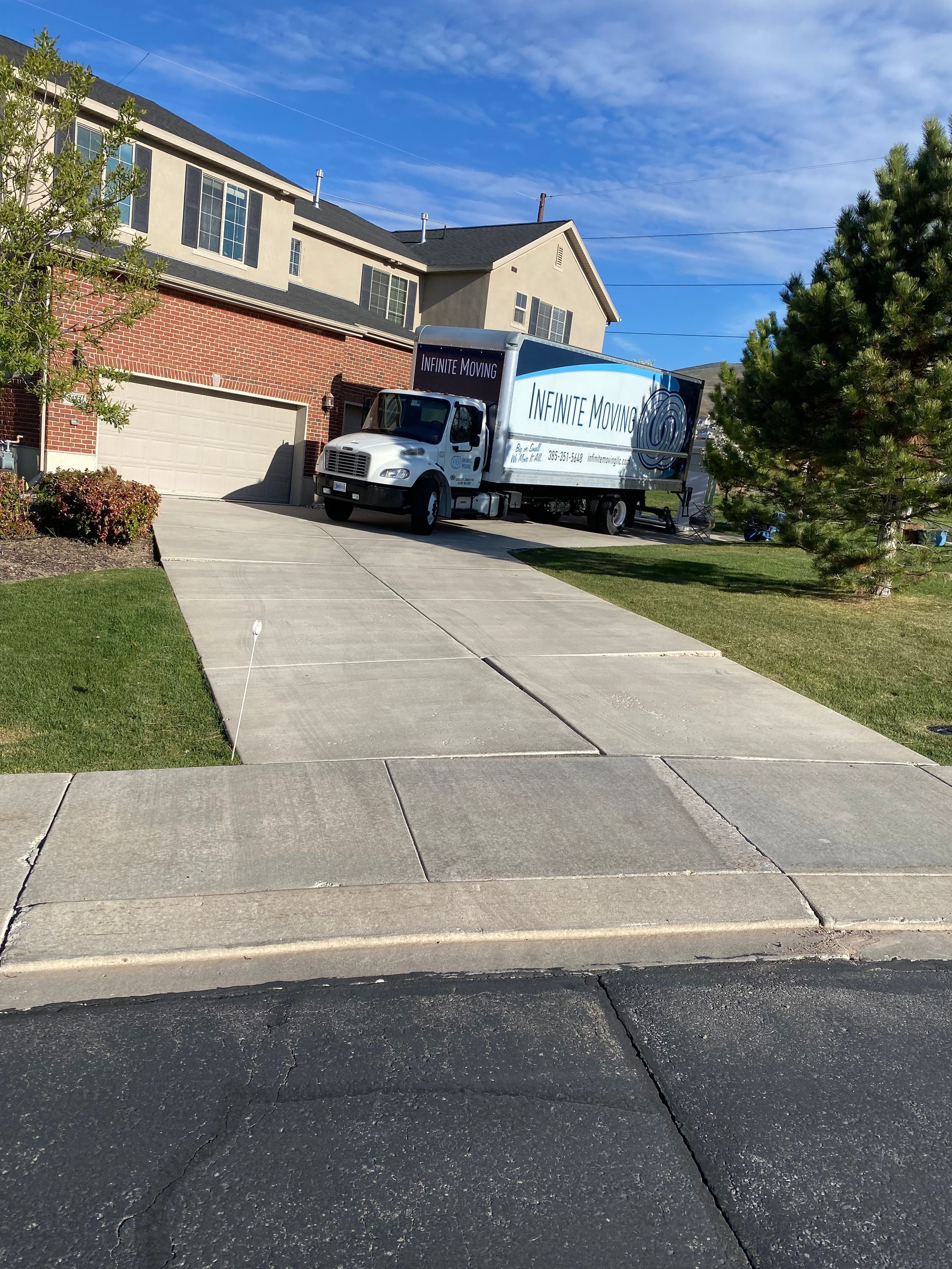 A white moving truck parked in a driveway, with a white box truck nearby, against a suburban house on a sunny day.