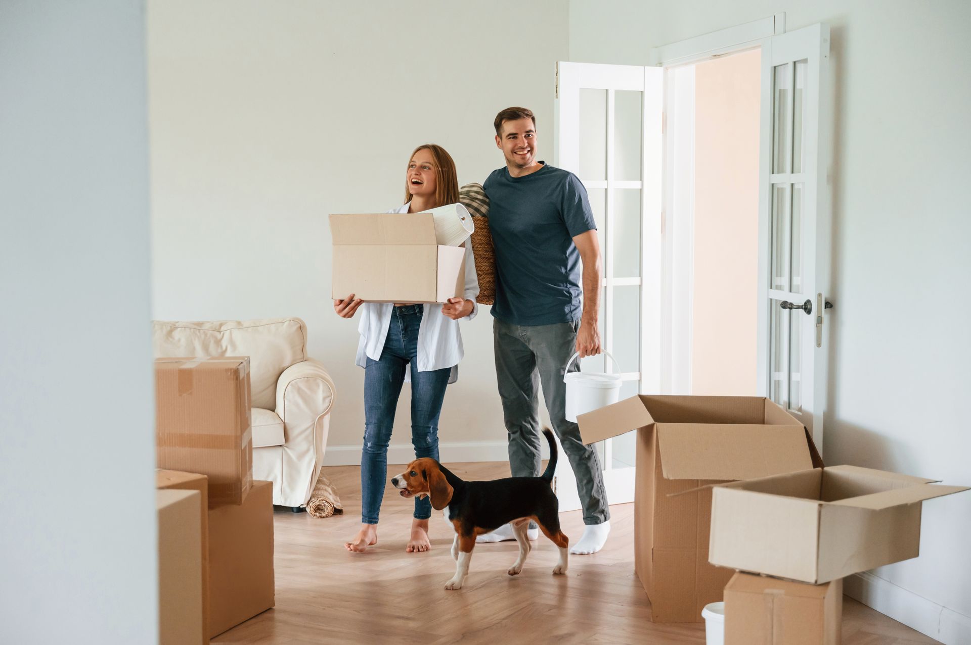 Couple holding a box, moving into a new home with their dog; boxes surround them.