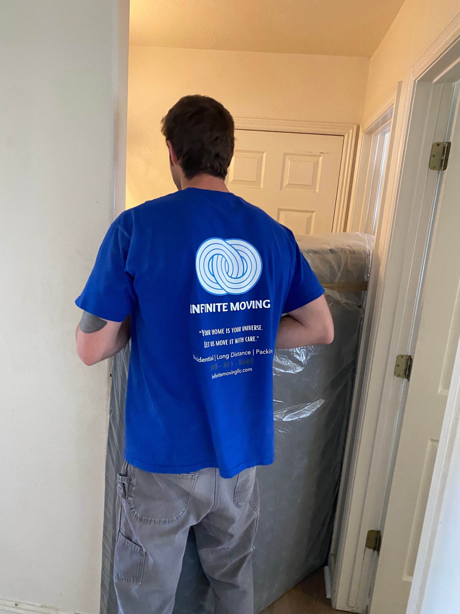 Man in blue shirt carrying a wrapped mattress through a doorway.