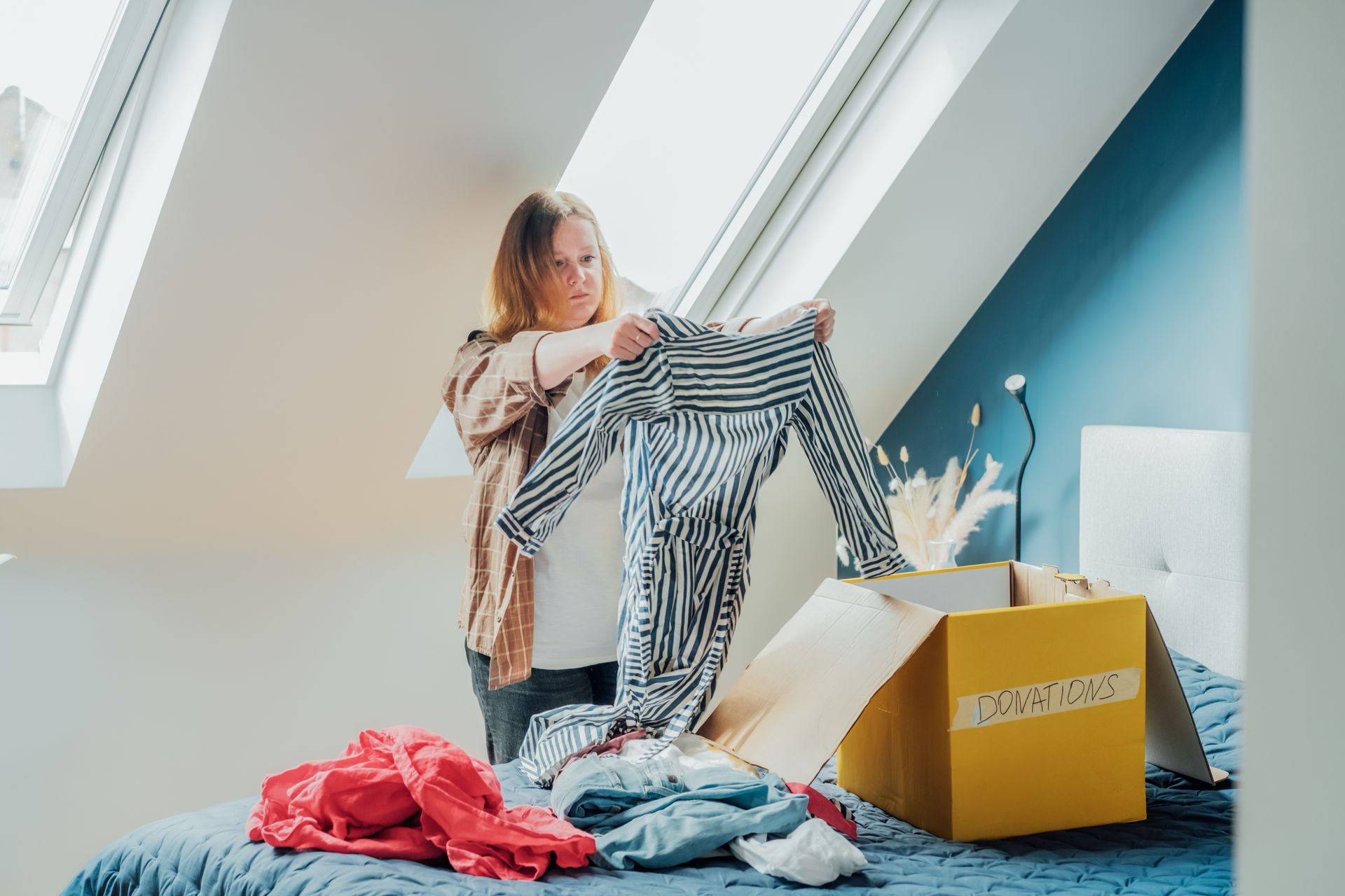 Woman sorting clothes from a box on a bed in a room with a sloped ceiling and window.