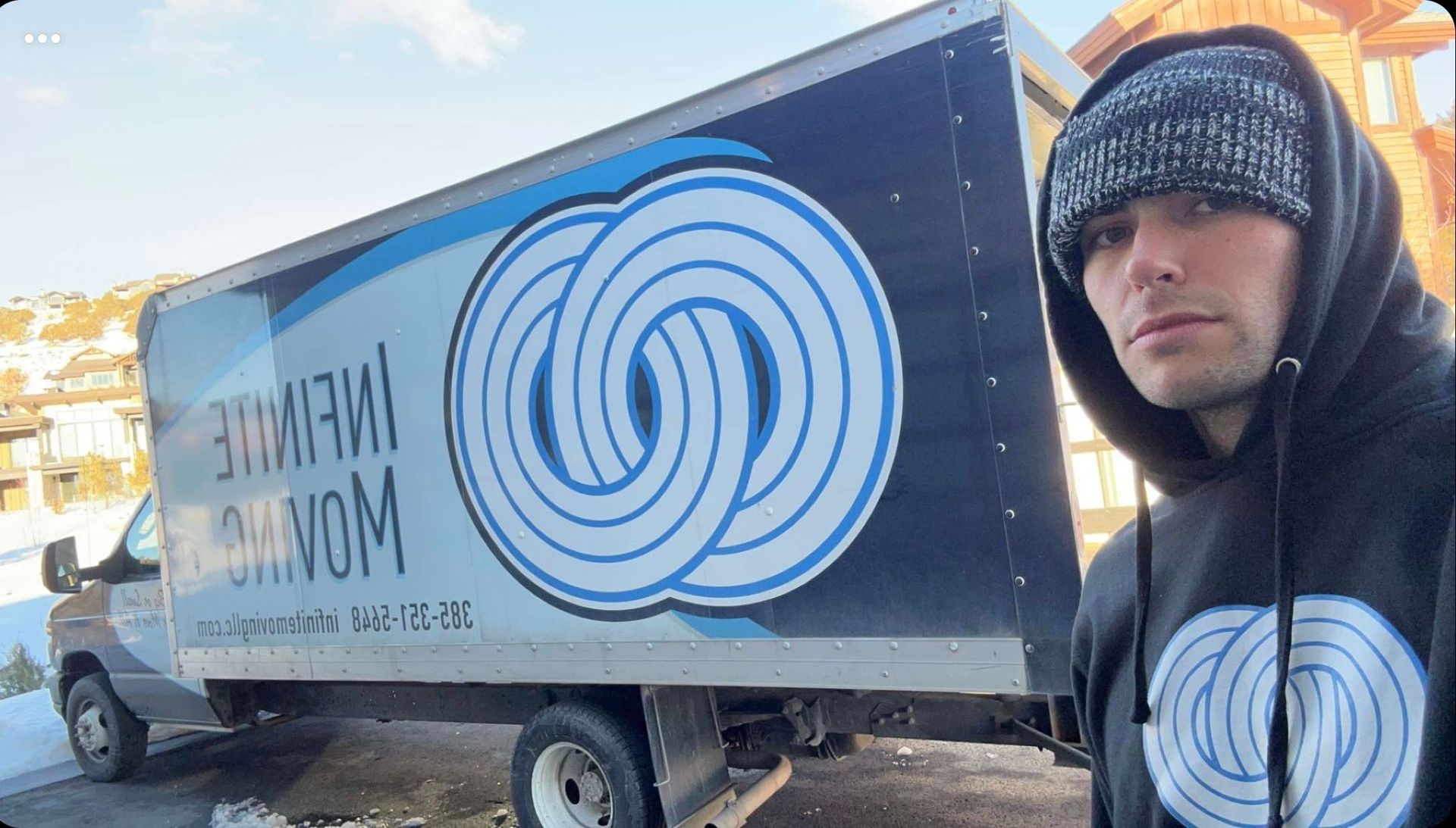 Man in beanie and hoodie standing next to a moving truck. Truck says 