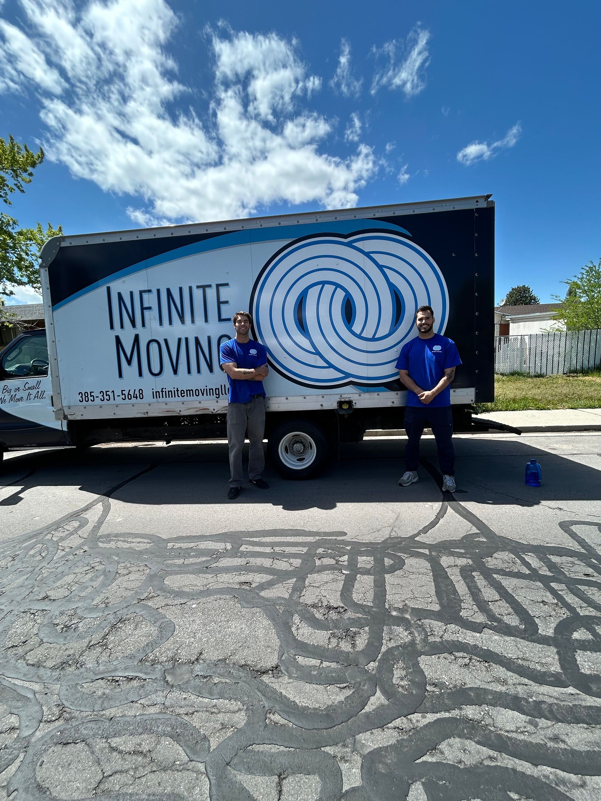Two men stand by an Infinite Moving truck on a street. Blue shirts, cloudy sky.