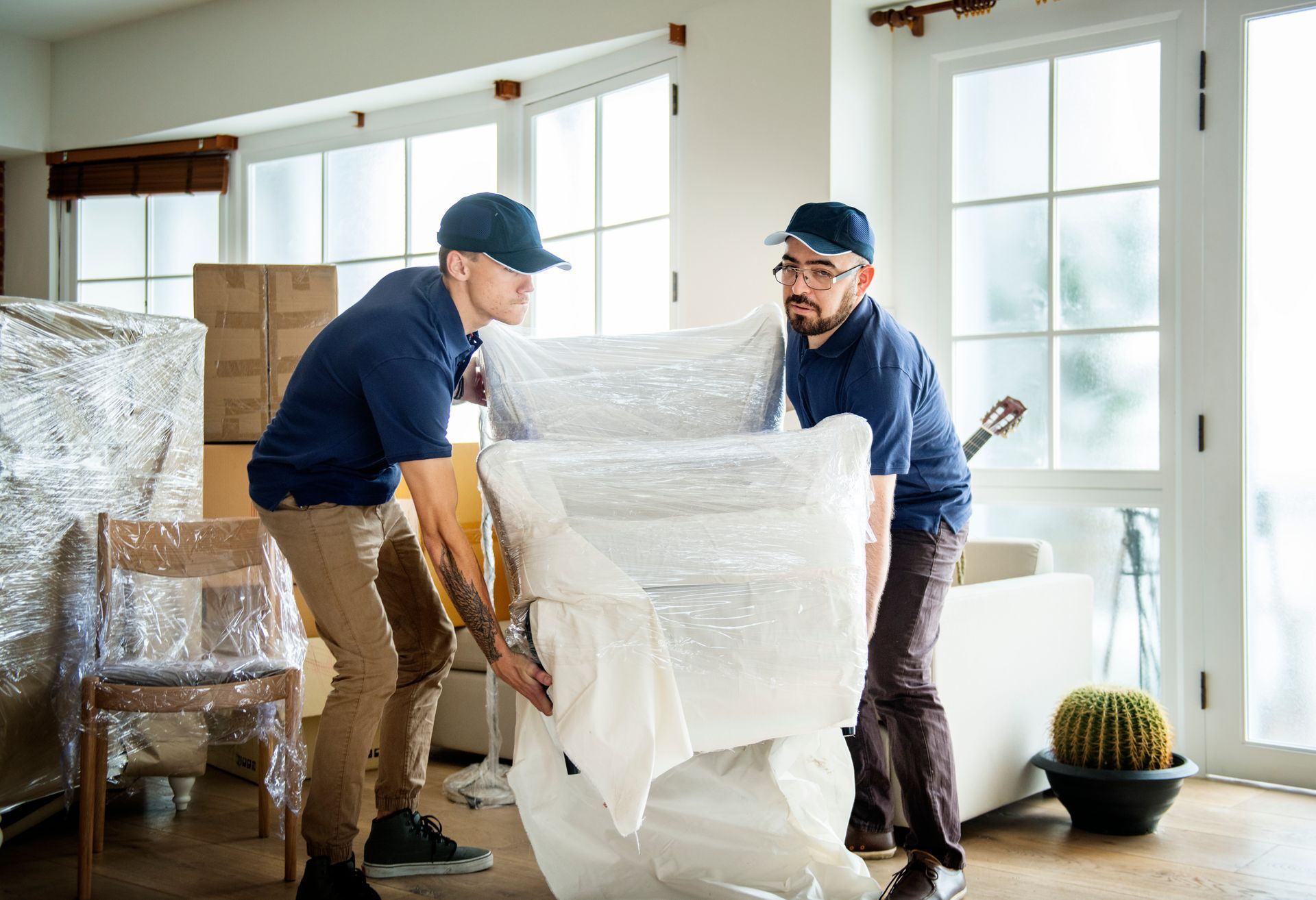 two men are carrying a couch wrapped in plastic in a living room .