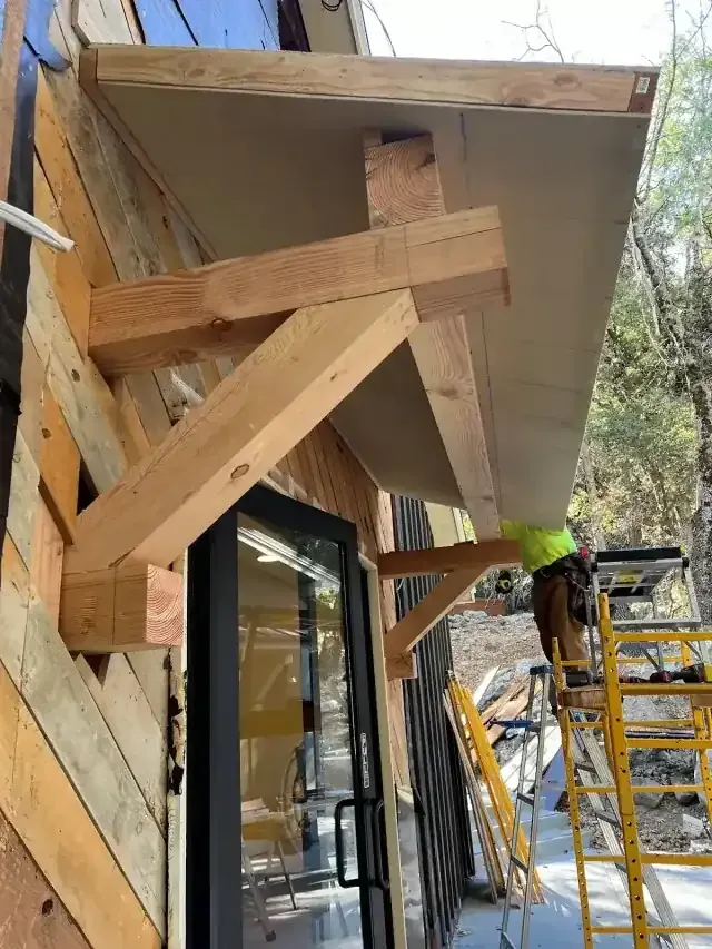 Wooden support brackets under a building overhang beside a window, with a ladder and worker in a hard hat.