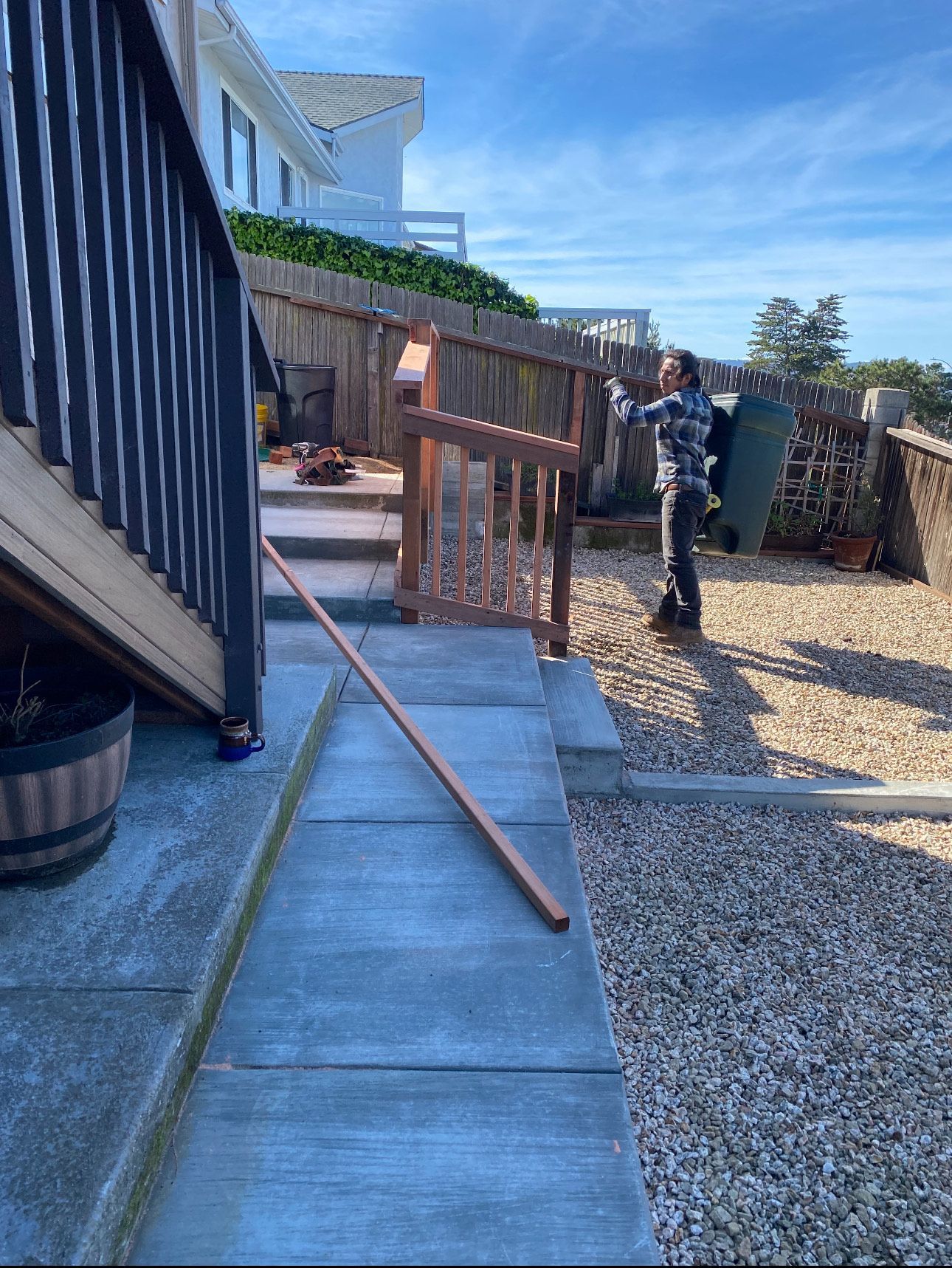 Person working on a wooden railing, outdoor setting. Steps, pebble ground, fence, and house in background.
