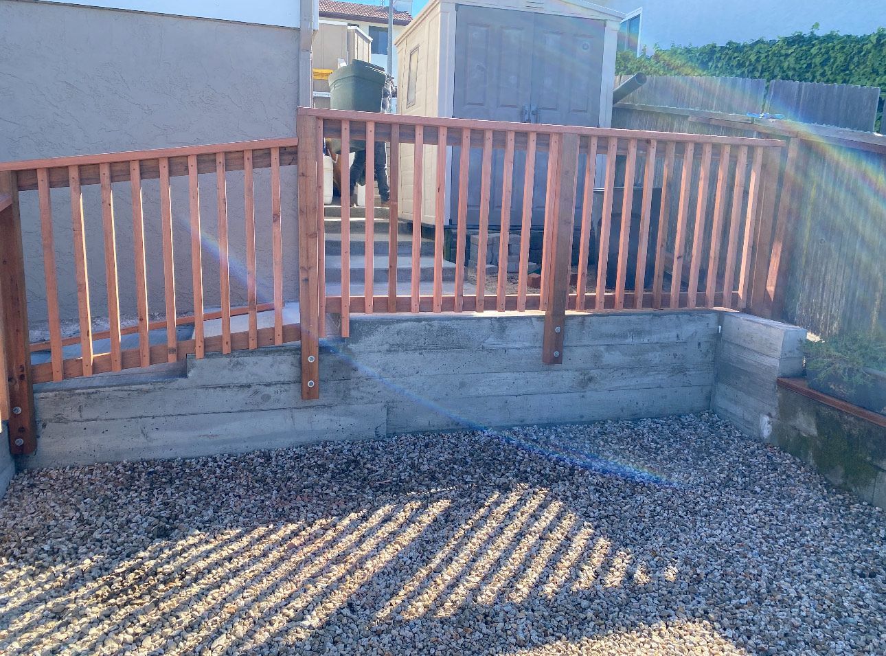 Wooden railing surrounds a concrete patio. Pebbles on the ground.