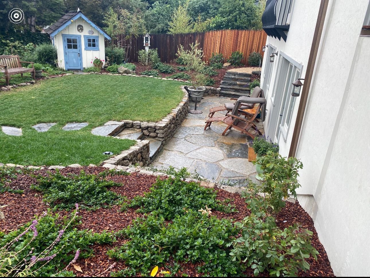 A backyard with a small blue shed, stone patio, steps, and lush green grass.
