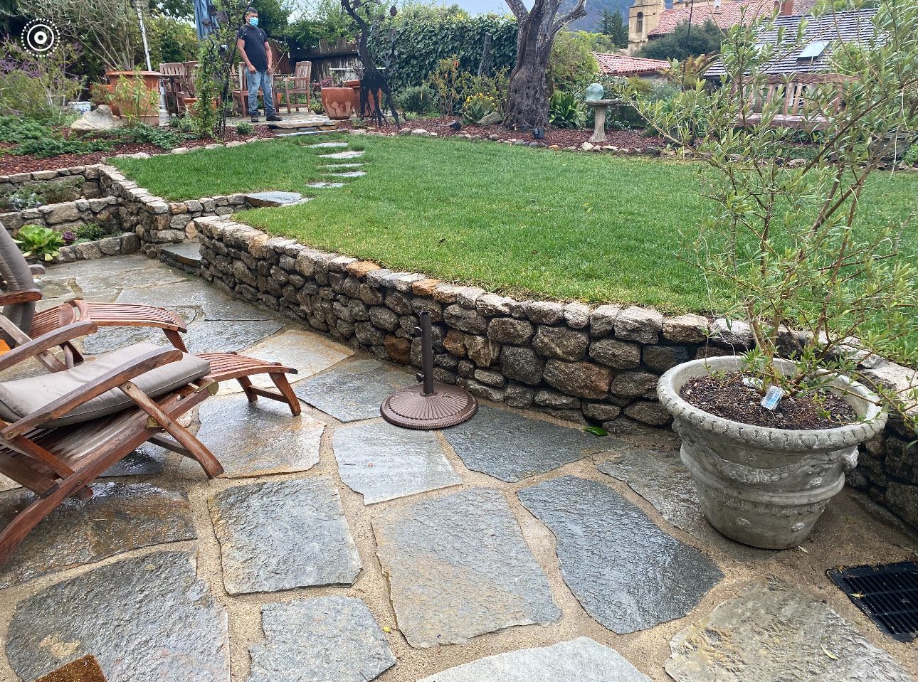 Stone patio with chair, stone wall, and small lawn area. Man in background.