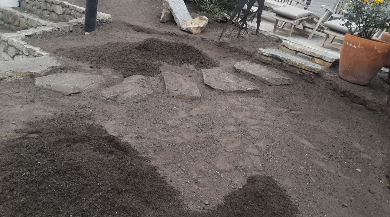 A partially constructed stone pathway with scattered stones on dark soil in an outdoor setting.