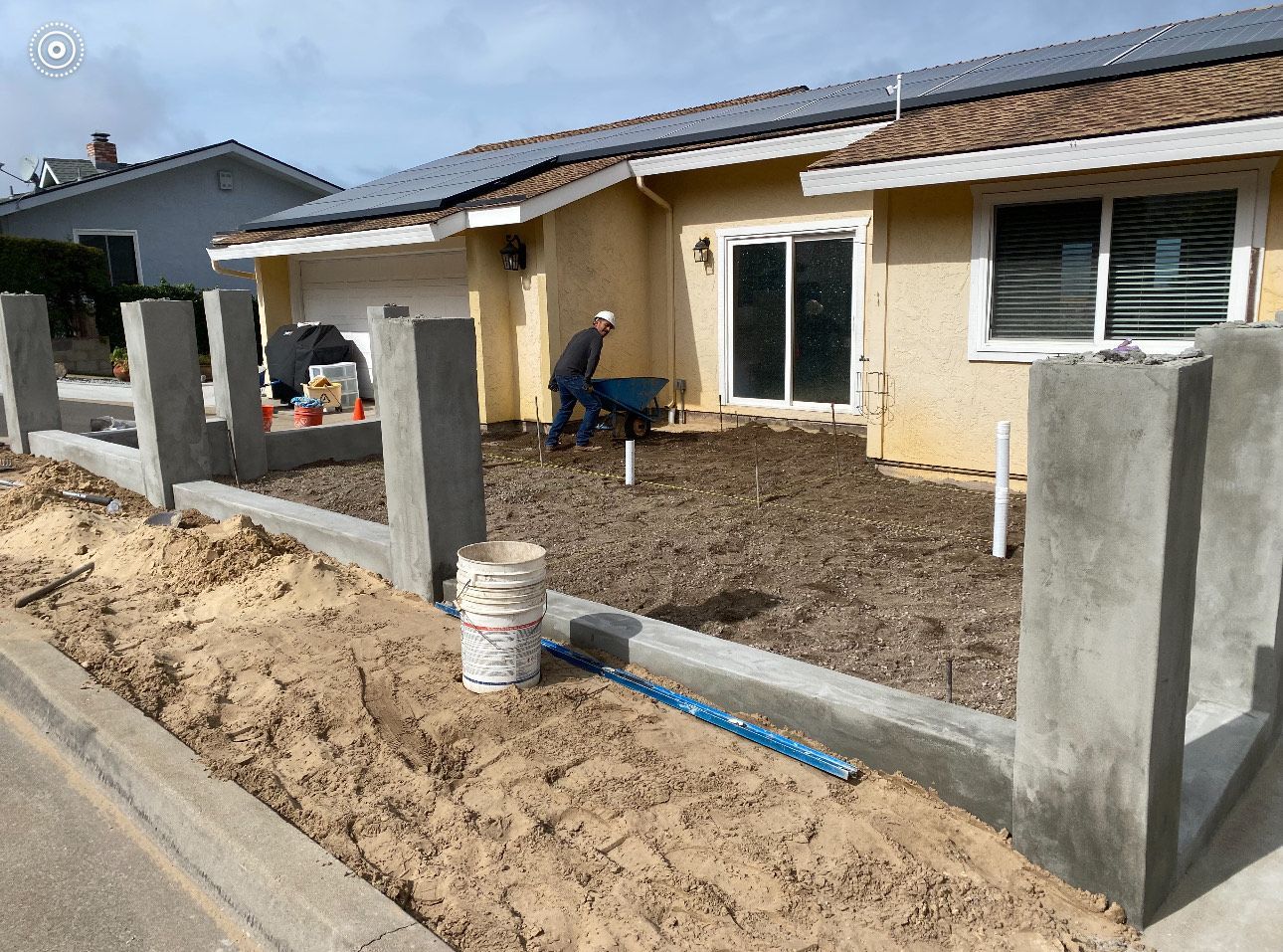 Construction of a concrete fence in front of a house; a worker uses a wheelbarrow.