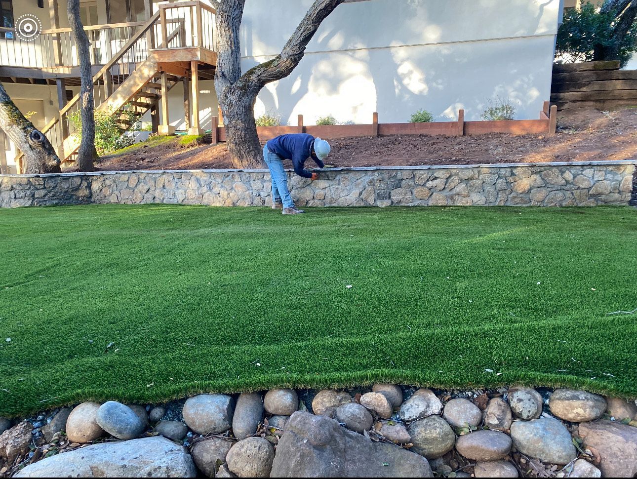 A person tending a wall with rocks, grass, and a tree in a backyard.