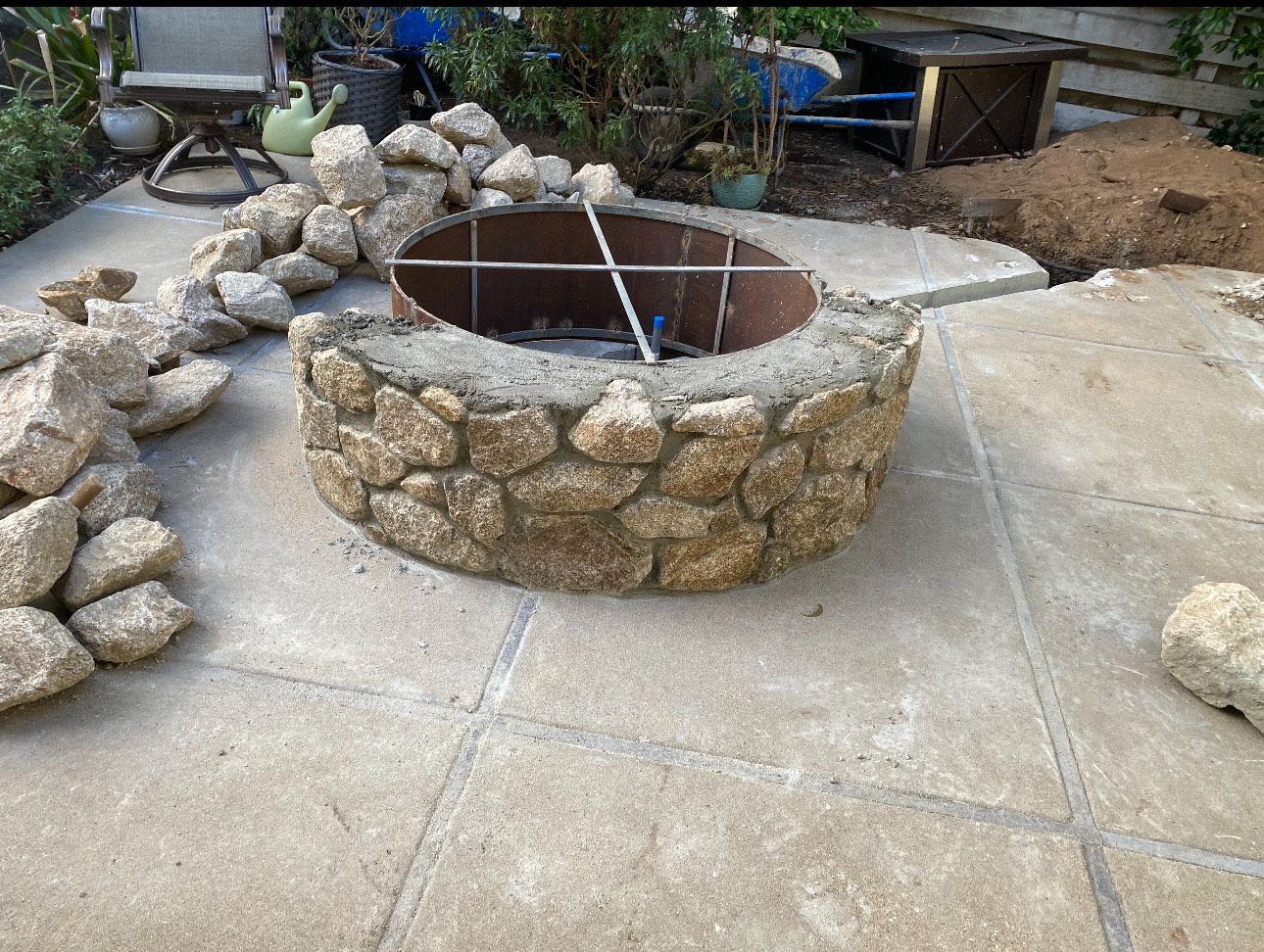 Fire pit made of stones, on concrete patio. Rocks piled nearby.