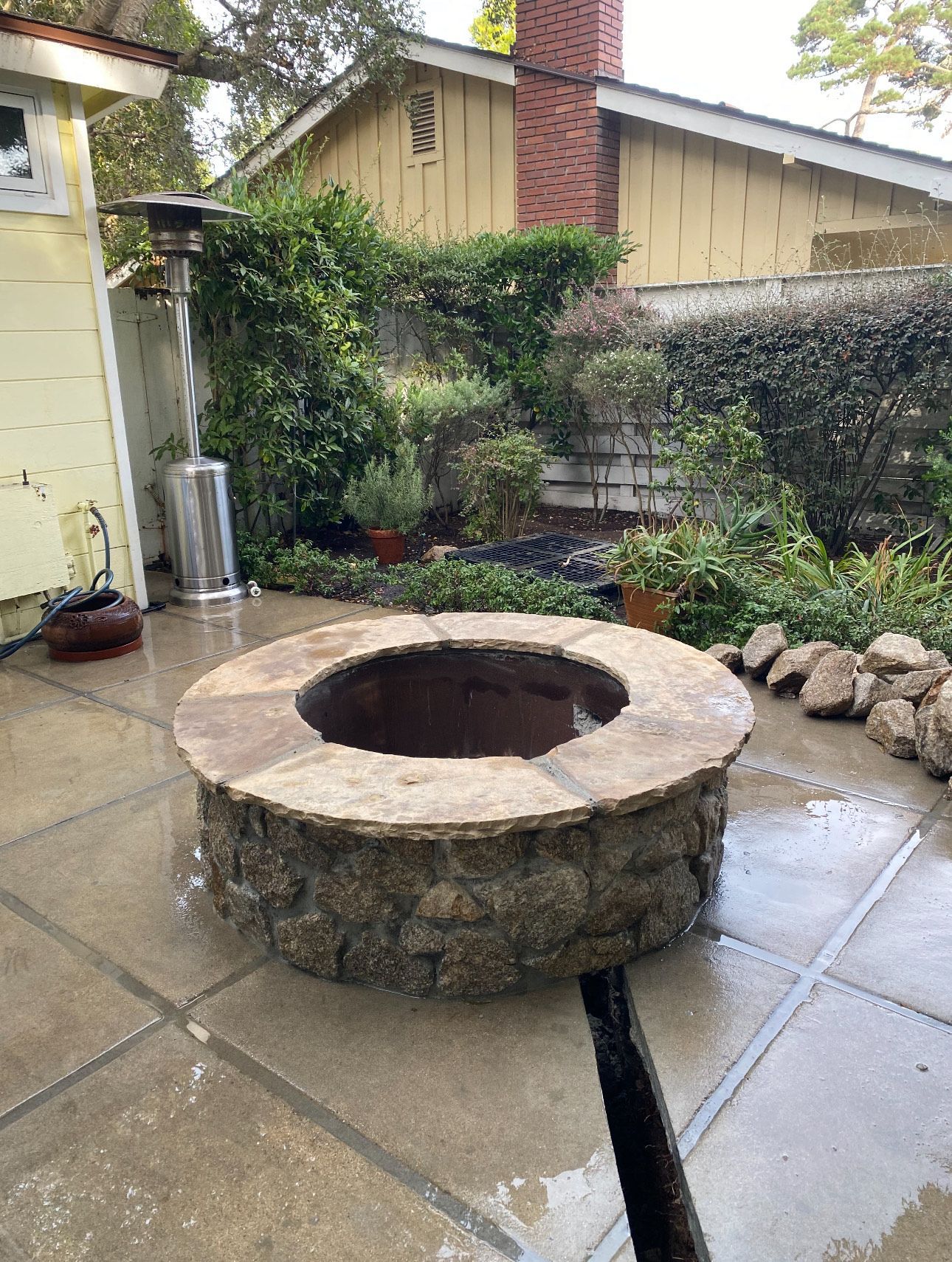 Stone fire pit in a backyard with a heater and lush greenery.