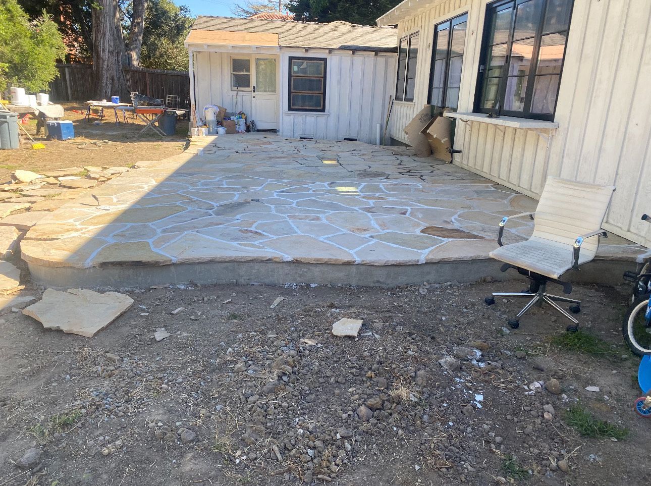 Backyard patio with flagstone and a small shed.  White siding, chair, and gravel ground.