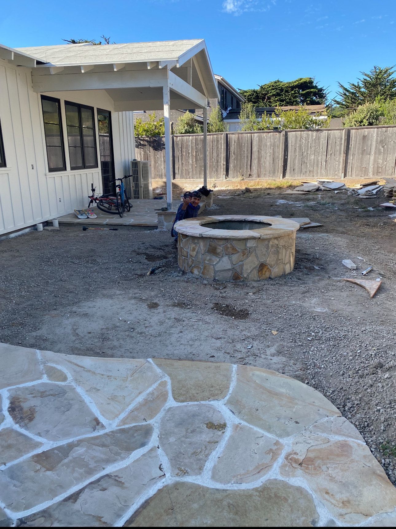 Backyard with a stone fire pit and patio, a person working on it, and a white house.
