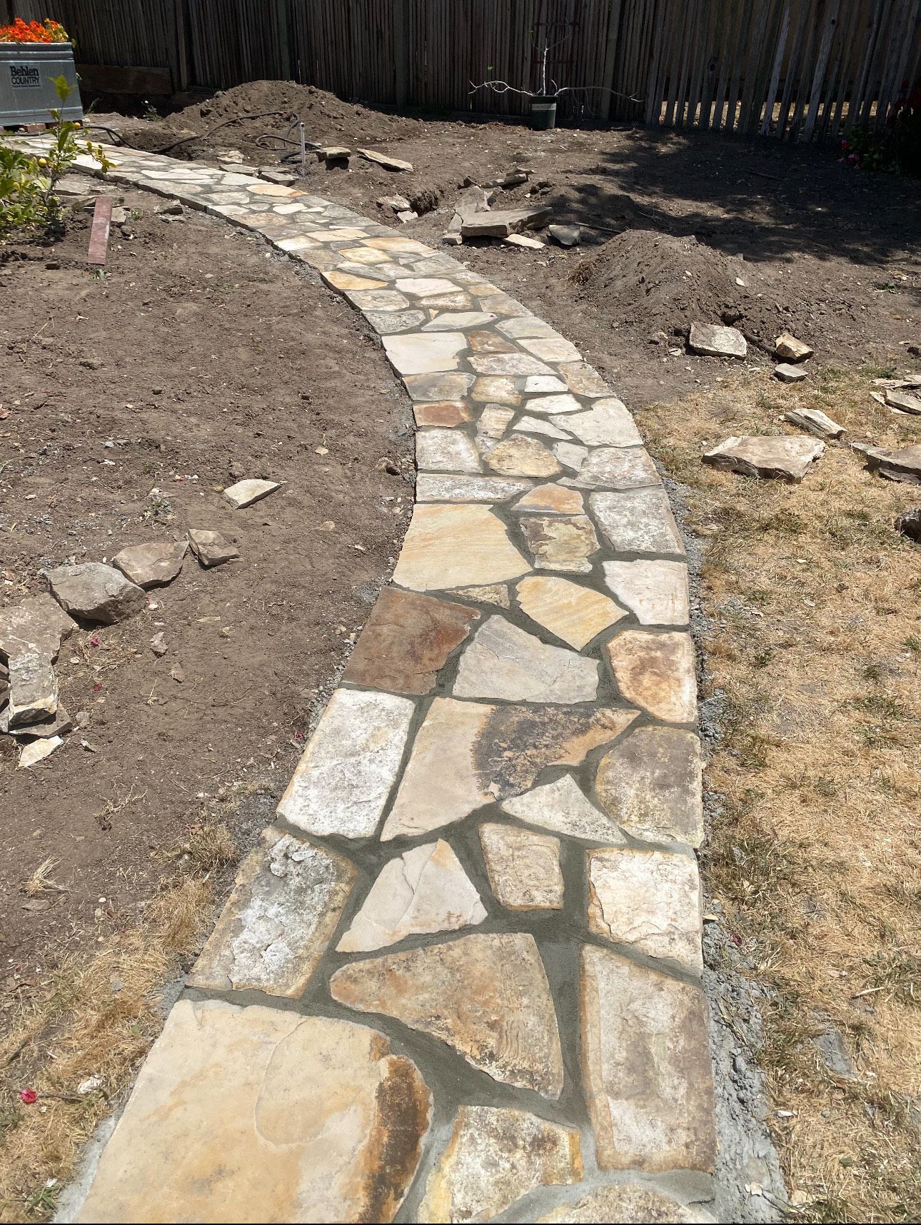 Stone pathway curving through a yard, surrounded by soil and grass, with a wooden fence in the background.