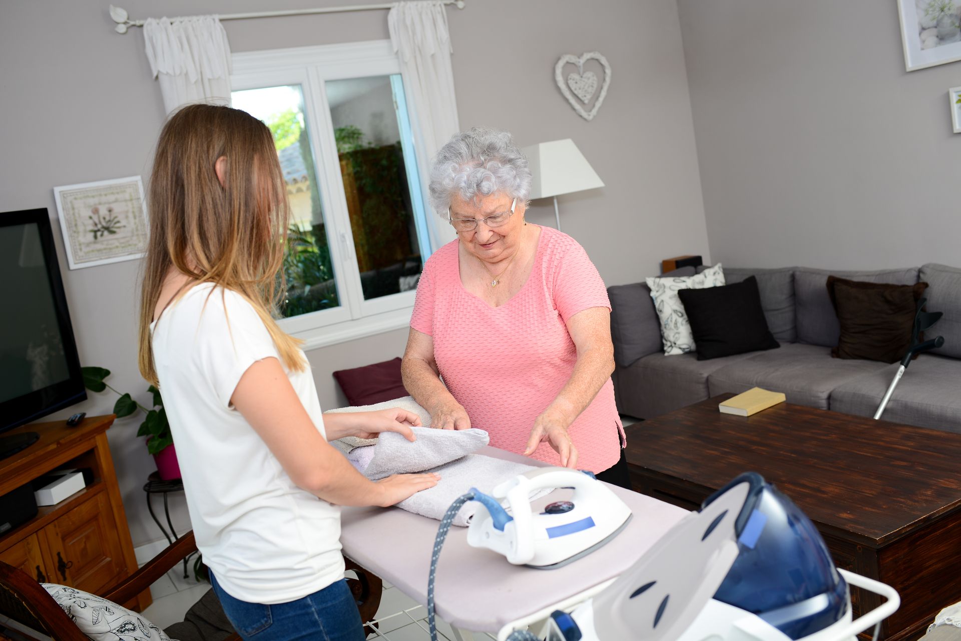 Woman ironing, assisting older woman. In living room, light from window.