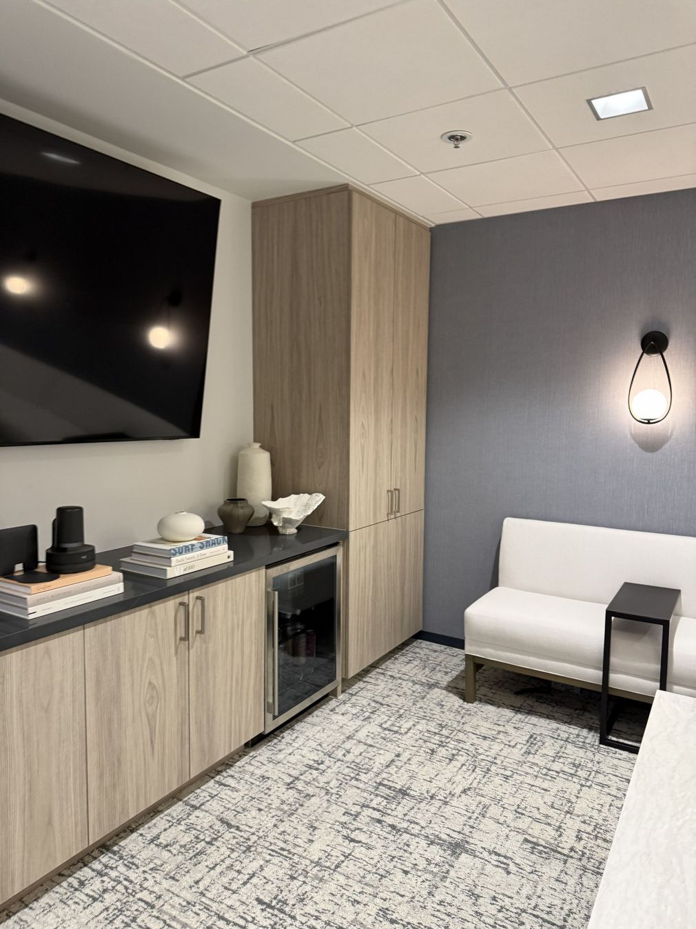 Modern office lounge with wall-mounted TV, wood cabinets, sofa, coffee table, and patterned carpet