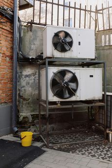 Two air conditioners are stacked on top of each other next to a yellow bucket.