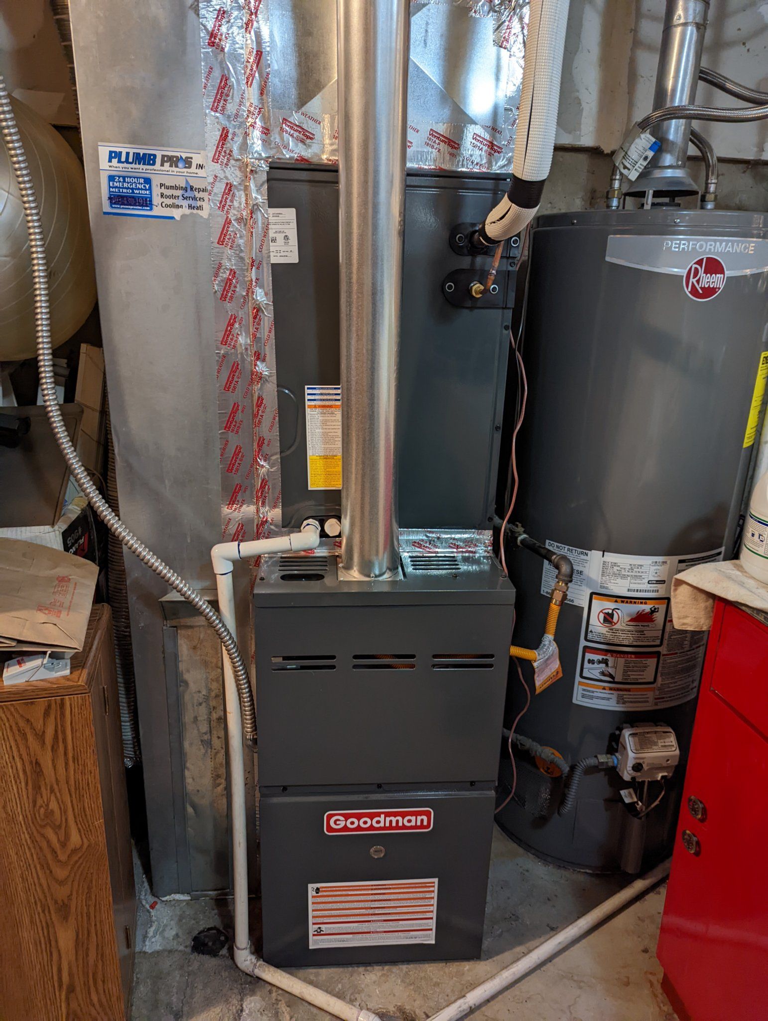A Goodman furnace and water heater in a utility room. Gray appliances with silver exhaust pipes.