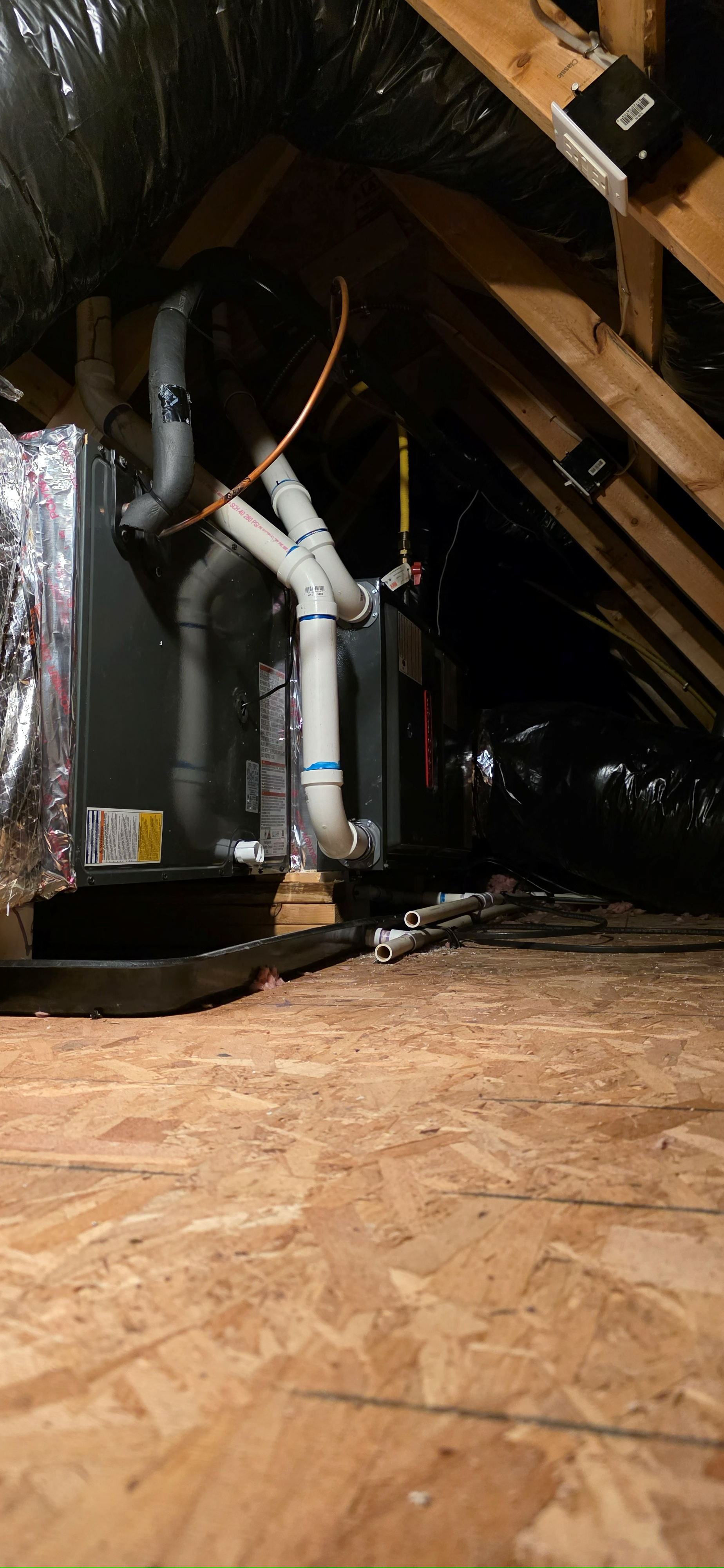 HVAC system in an attic. Wooden beams and plywood flooring. Black insulation and gray pipes.