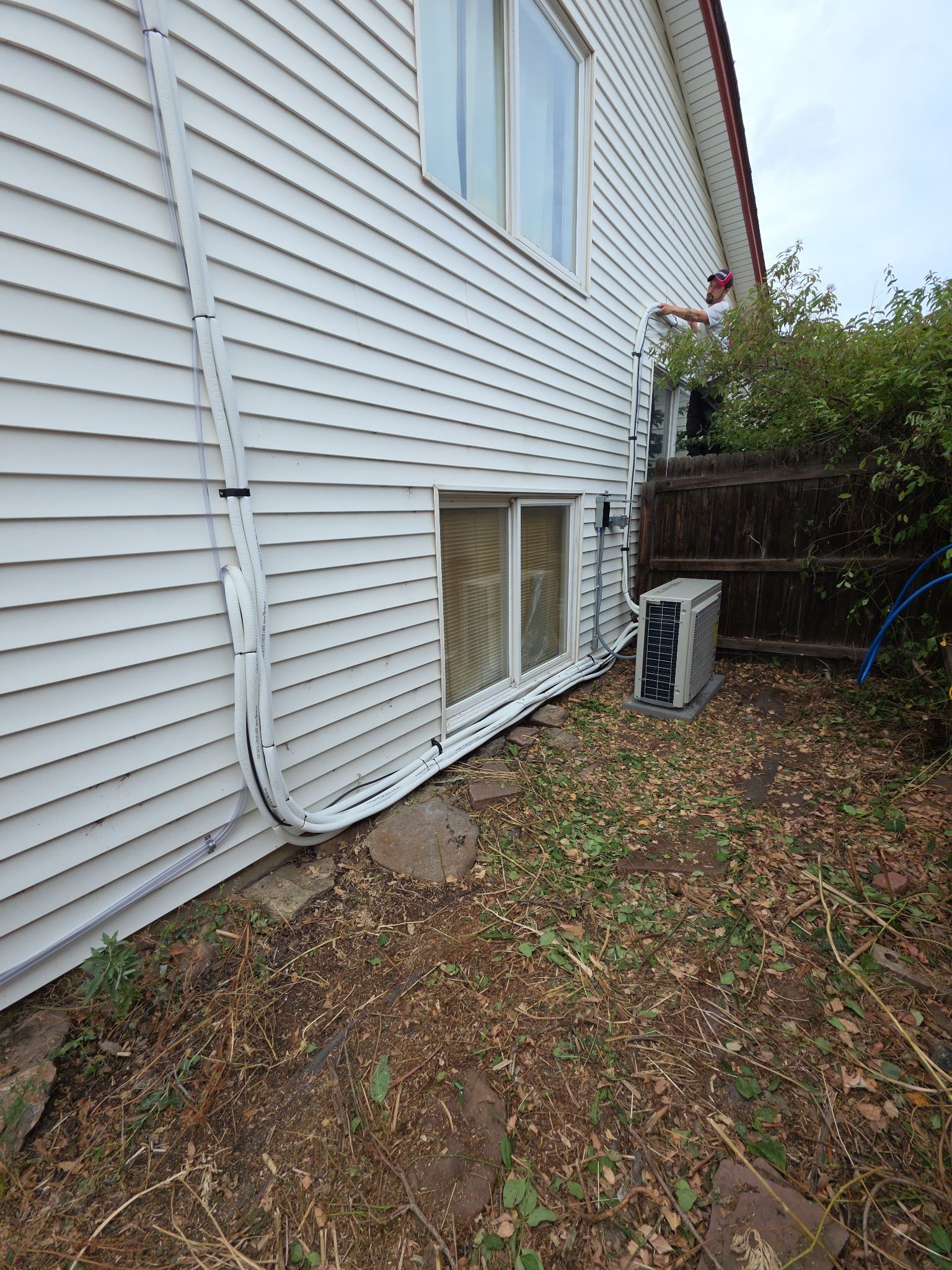White siding of a house with an air conditioner unit outside. Piping runs along the wall and ground.