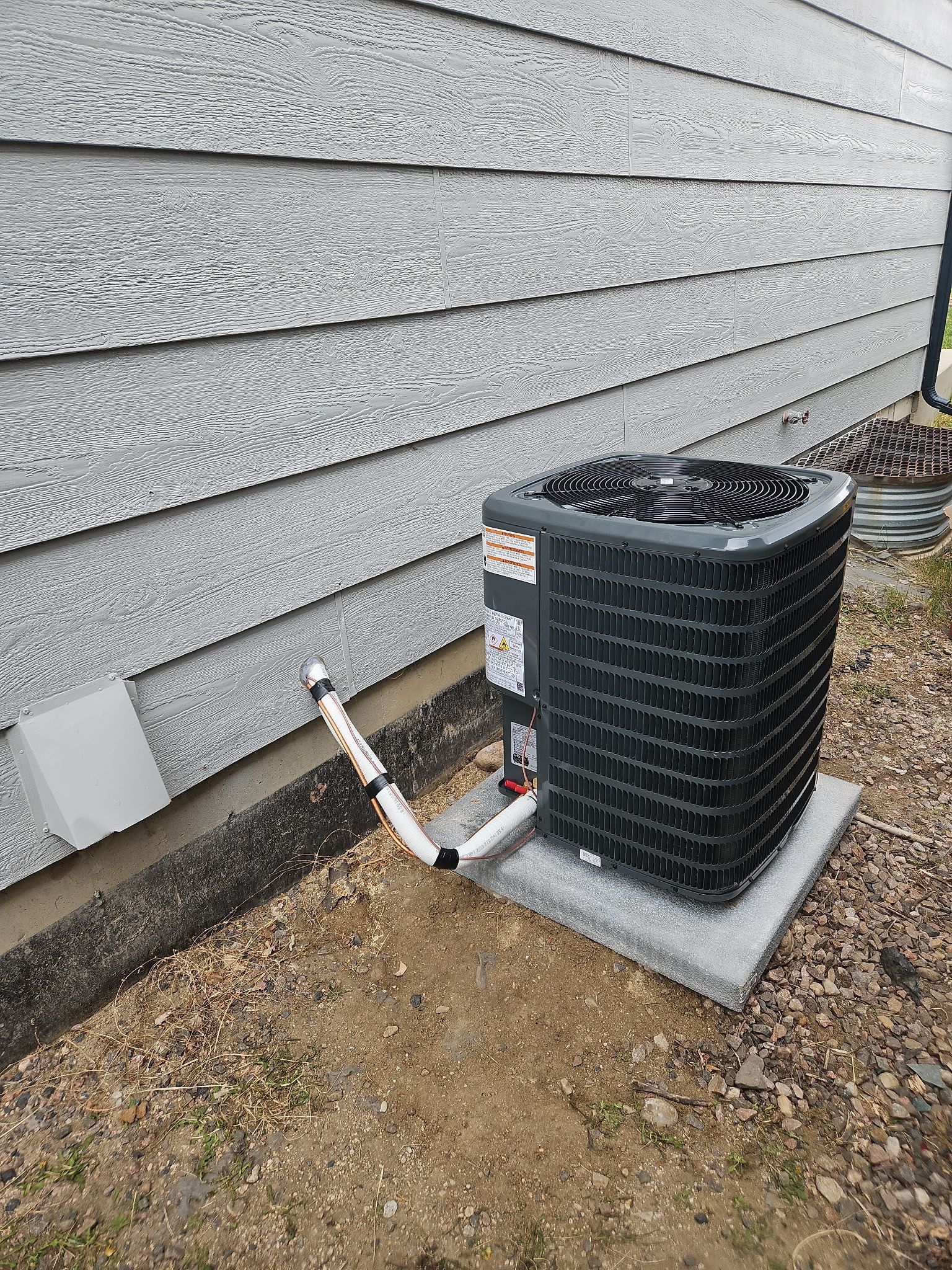HVAC unit next to a building with gray siding, pipes and a concrete base, set in a yard.