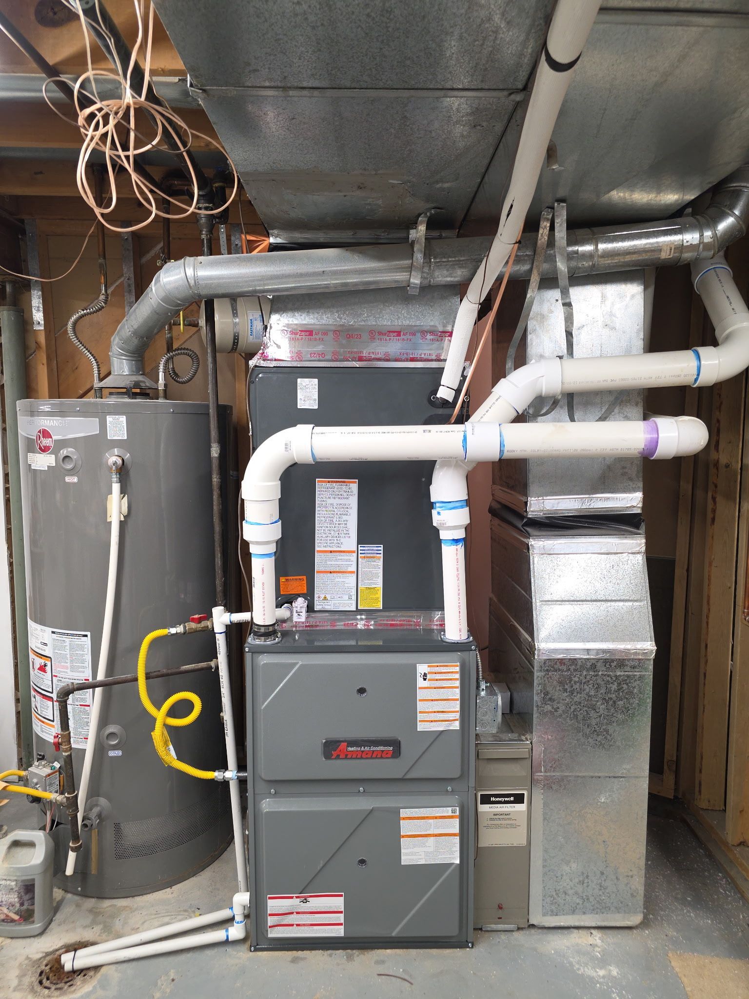 Furnace, water heater, and ductwork in a utility room. Gray appliances with white PVC pipes and metal ducting.